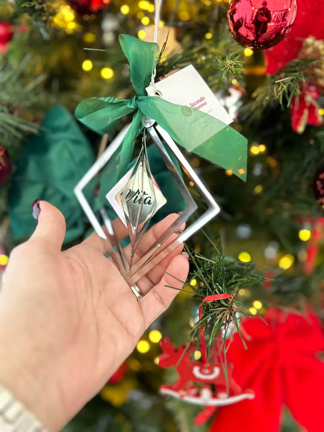 L'addobbo in acciaio del Natale 2024: due rombi sospesi e al centro la scritta «Vita», legato con un nastro verde e il biglietto di Seconda Mamma, tenuto in mano davanti all'albero.